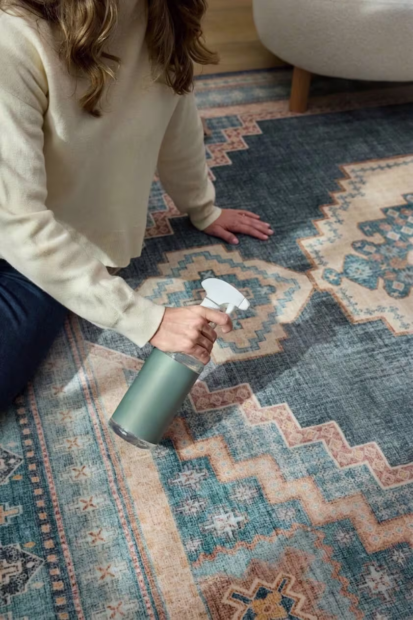 A close-up view of a bottle of spot cleaner being sprayed onto a traditional-style blue geometric rug.