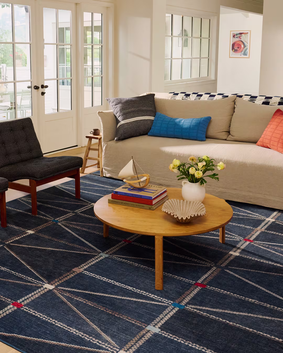 A living room with a beige sofa, colorful pillows, a round wooden coffee table with books and a vase of flowers, a dark chair, and large windows letting in natural light.