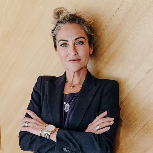 Person with hair in bun wearing navy blazer and jewelry, arms crossed, against light wooden background.
