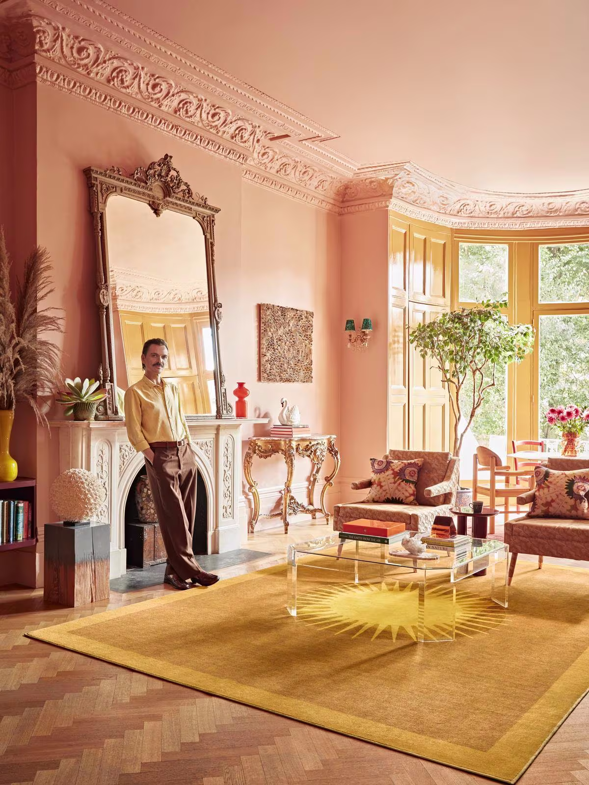 Image of a bright living room where interior designer Matthew Williamson stands with two leather chairs, a fireplace, and the Matthew Williamson Helios Gold Rug displayed. 