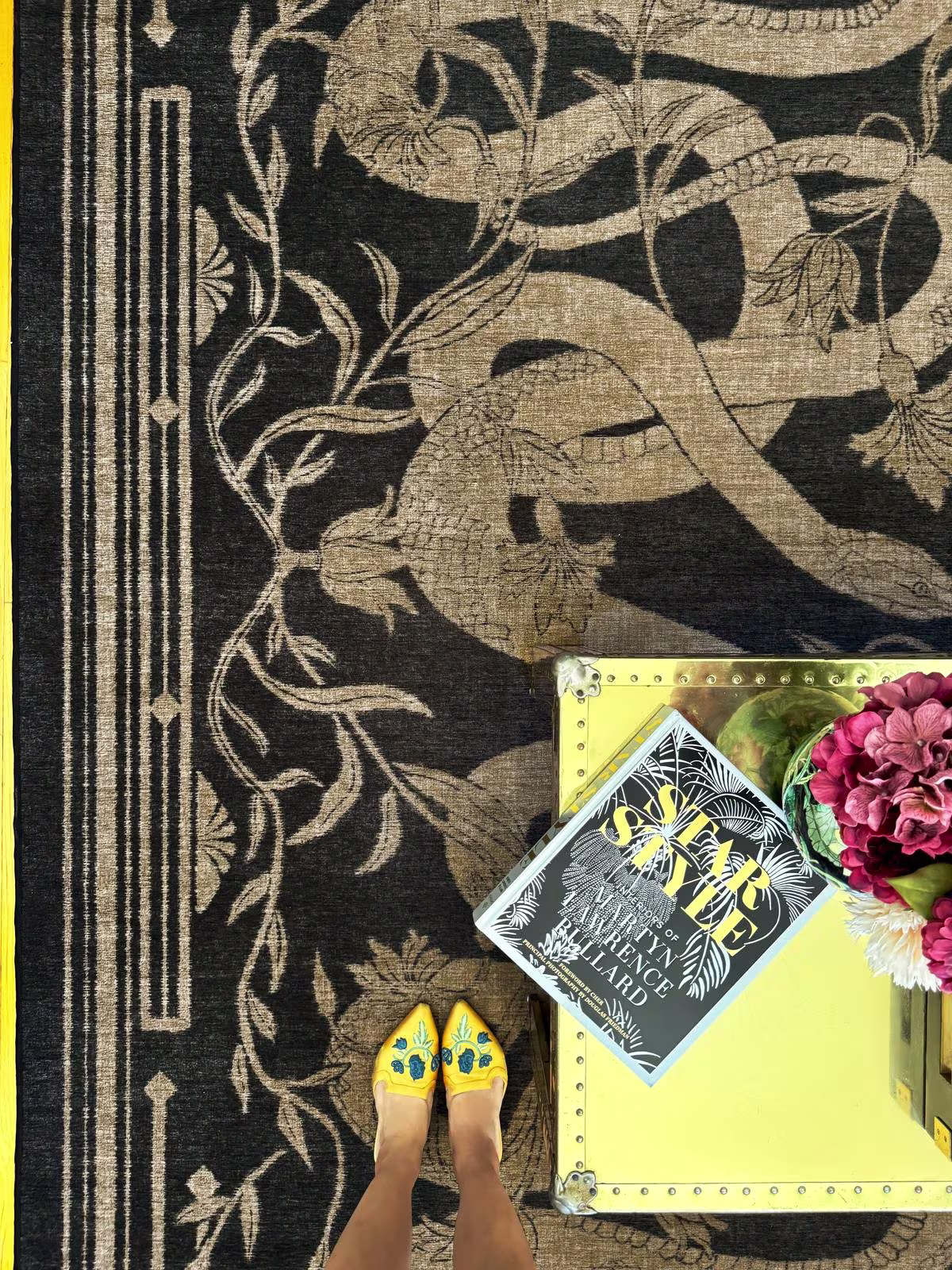 Ruggable x Martyn Lawrence Bullard Sumaya Soft Black Rug with gold trunk, bold floral arrangement, and yellow embroidered slides