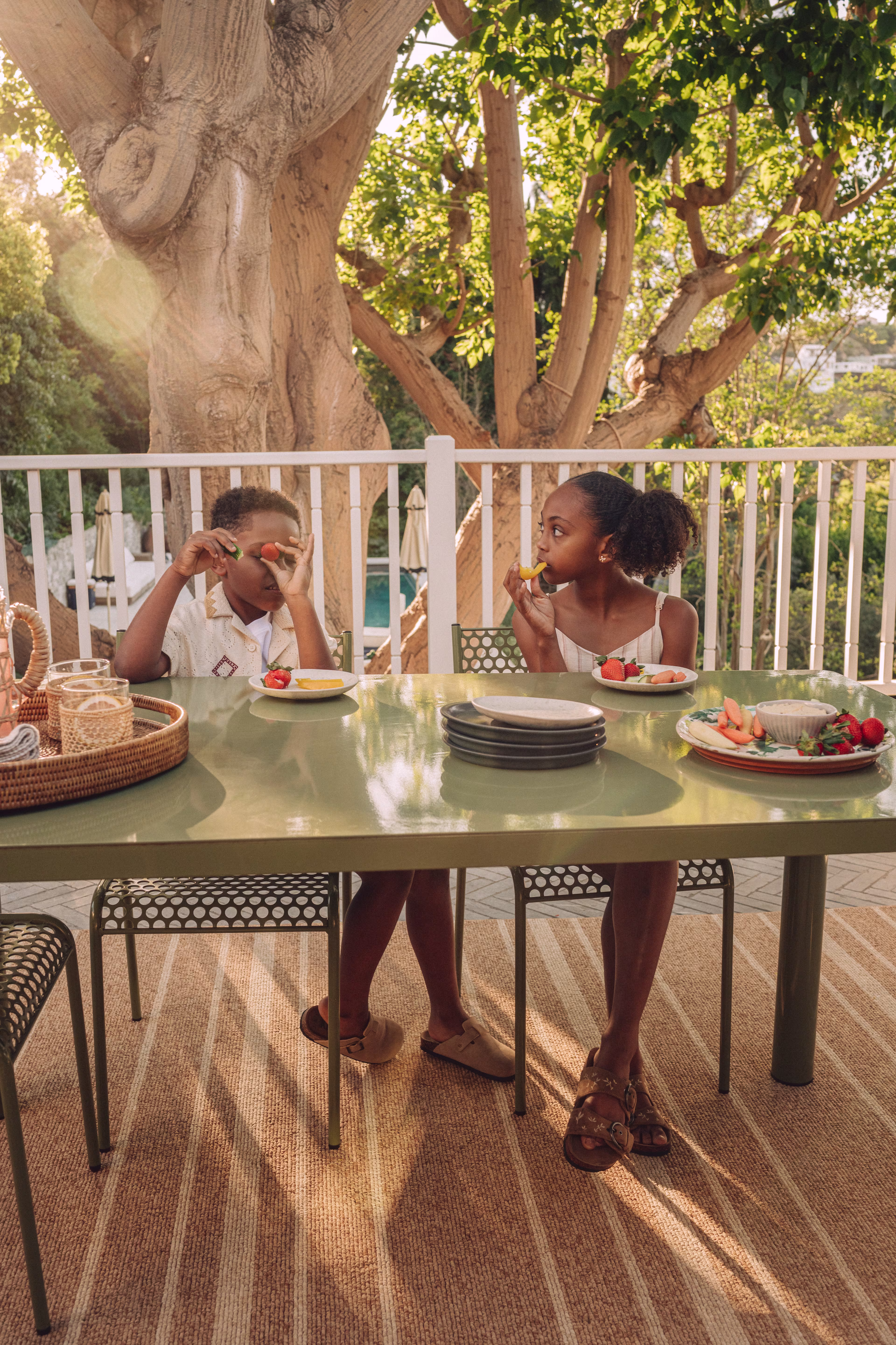Two children sit at an outdoor table on a sunlit patio, eating fruit, with a large tree and greenery in the background and a striped rug beneath the table.