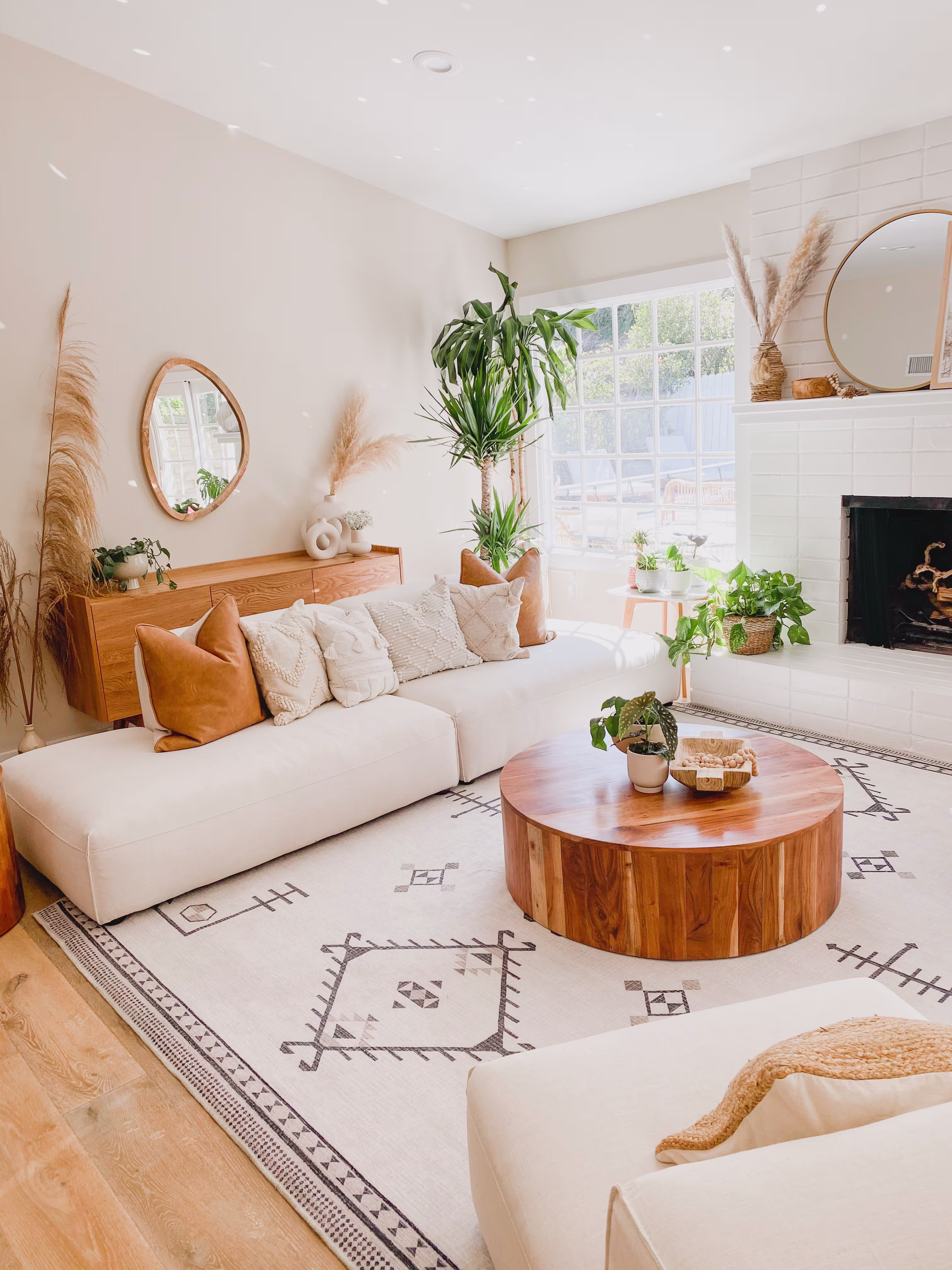 Bright living room with white walls, natural light, and a cozy atmosphere. Features a white sofa, wooden coffee table, fireplace, and indoor plants.
