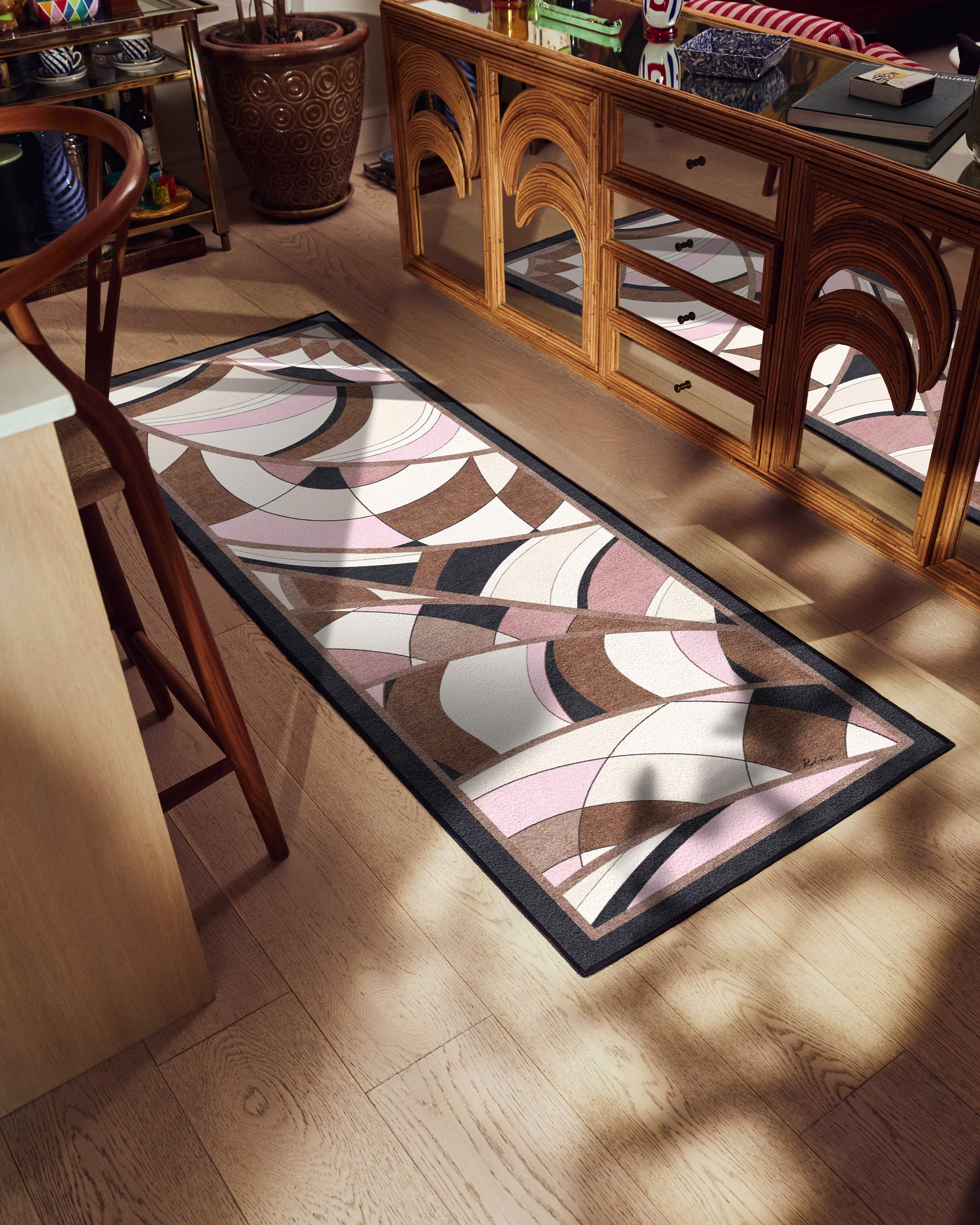 A long, abstract-patterned rug in pink, brown, and white tones lies on a wooden floor. Nearby, a mirrored cabinet reflects light, creating a warm, cozy ambiance.