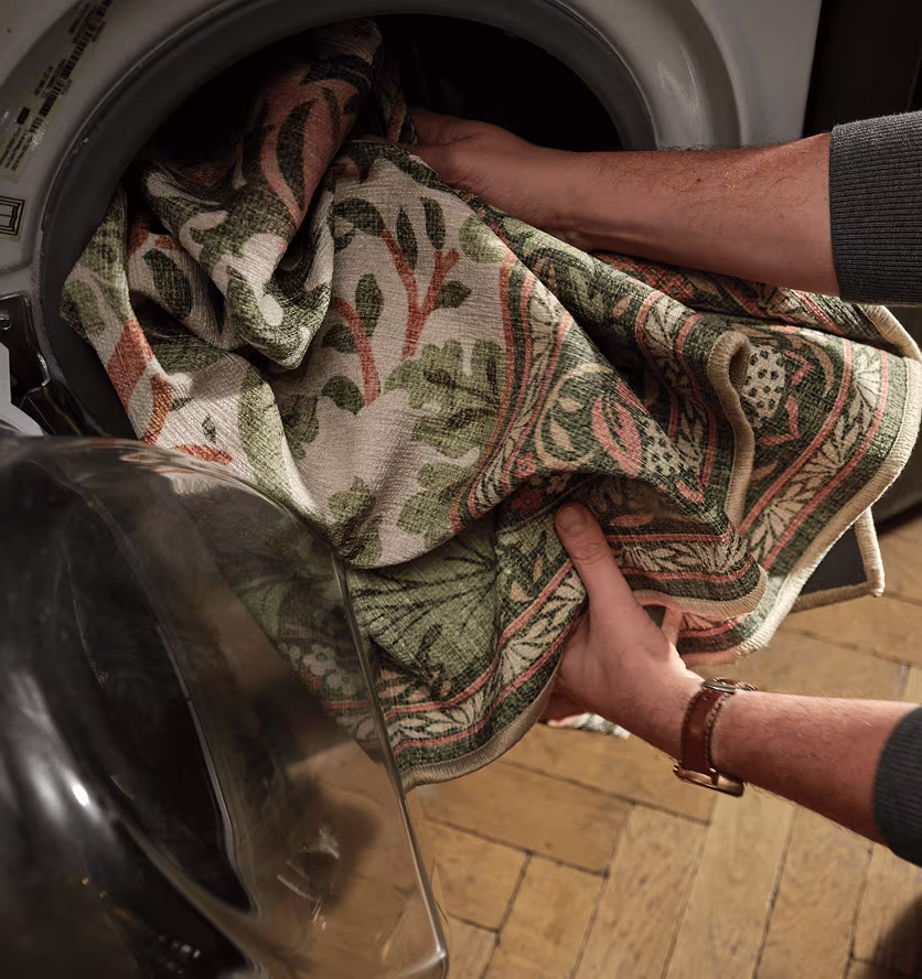 Colorful tree pattern rug in green, orange, and tan being loaded into a washing machine for a deep clean