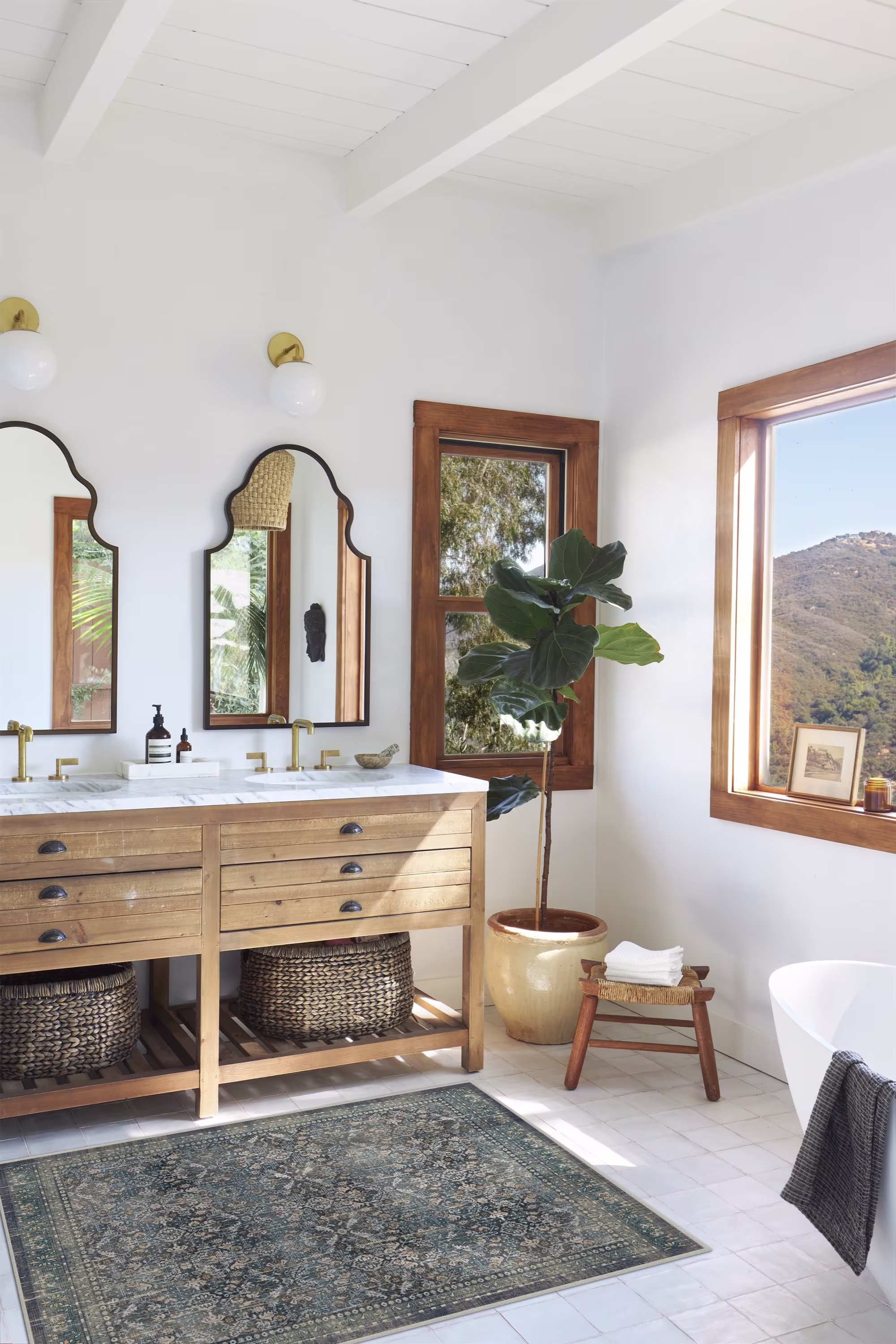 Ruggable Frances Vintage Teal Blue rug styled in a bright, rustic bathroom featuring a light wood double vanity with marble top, arched black-frame mirrors, and a tall fiddle-leaf fig tree.