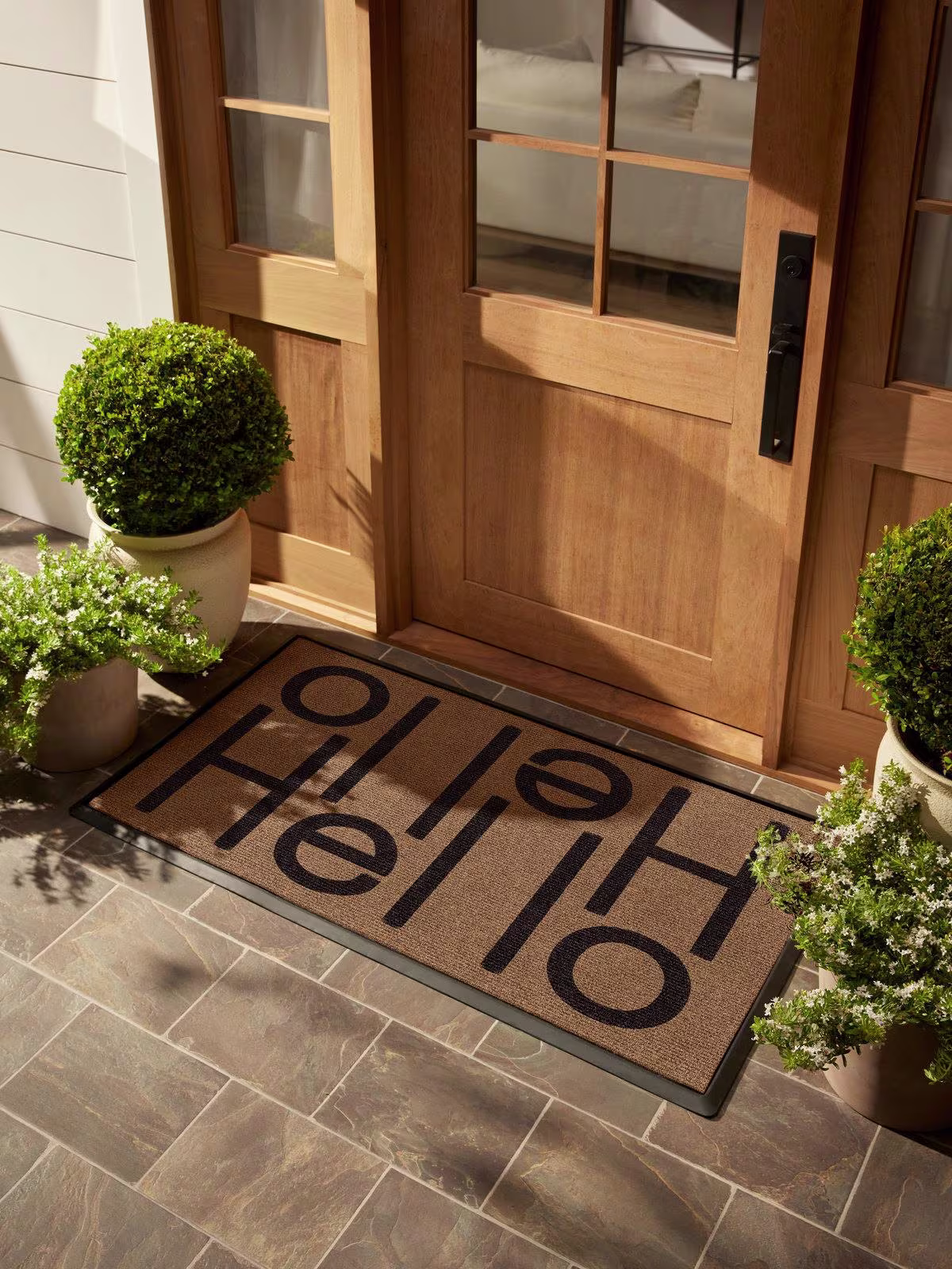 Image of a doorstep with plants and a wooden door featuring Ruggable's Hello Light Coir Estate Doormat. 
