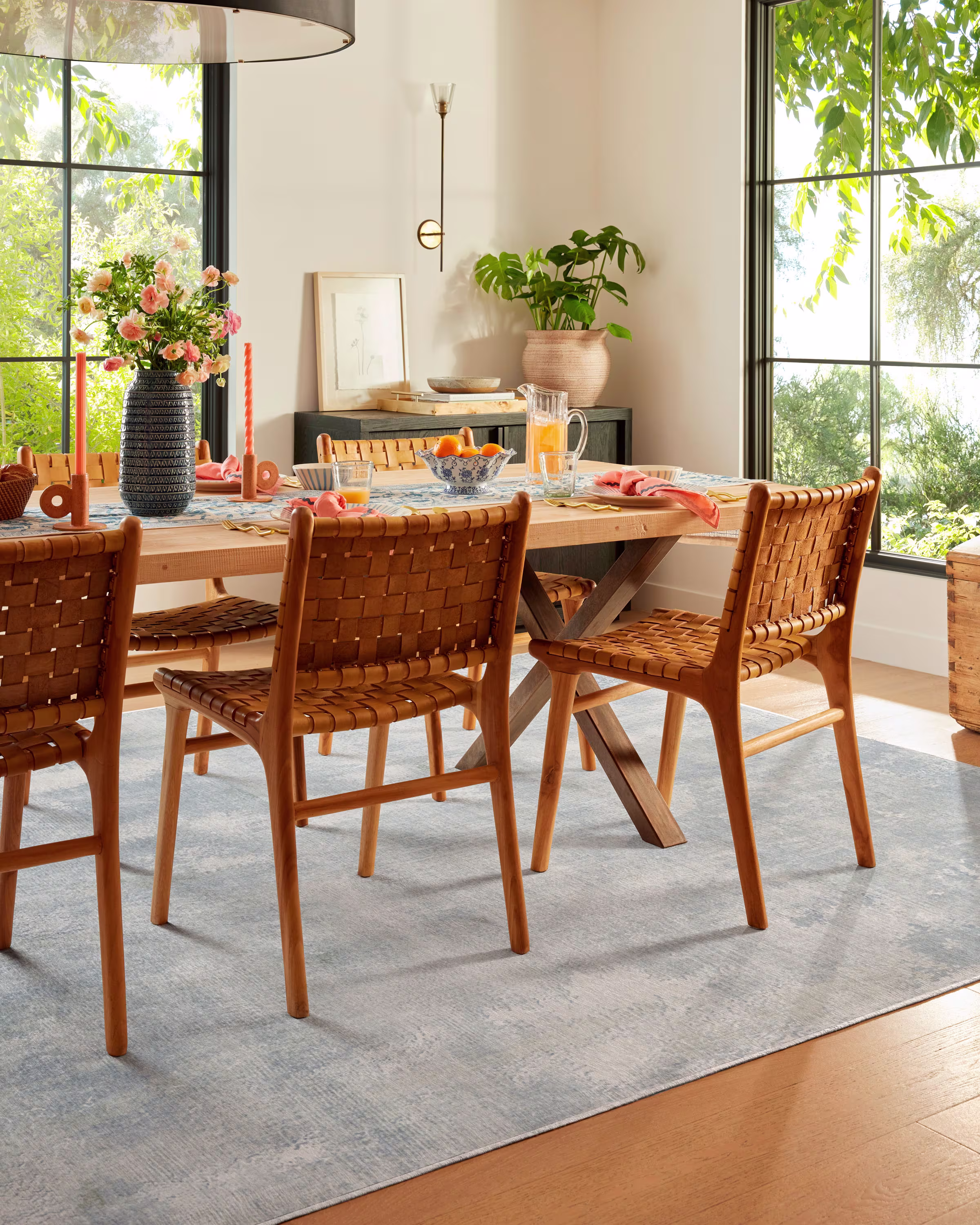 Bright dining room with a wooden table set for breakfast. Four woven chairs surround the table adorned with pink flowers and orange candles. Large windows overlook lush greenery, creating a serene, inviting atmosphere.