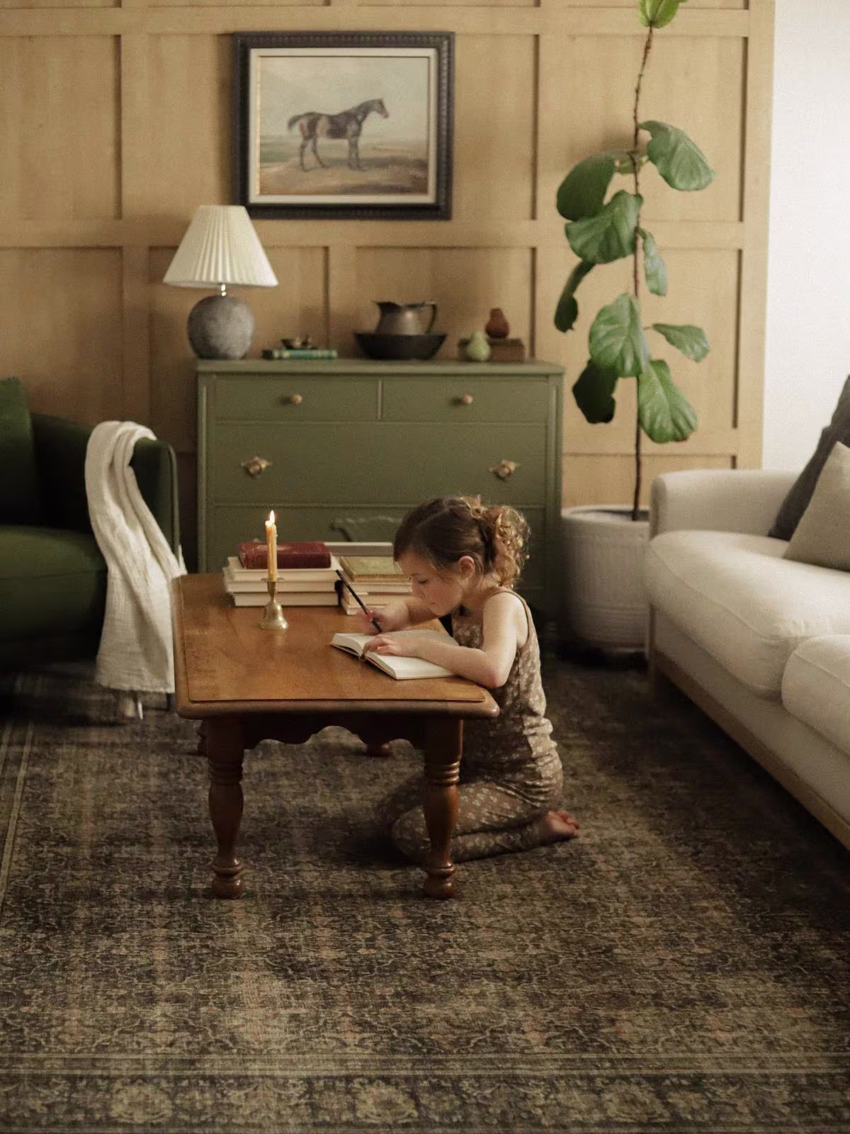 Ruggable Vesper Olive & Gold rug in living room with moody lighting, fiddle leaf tree, lit candle, and child writing in journal