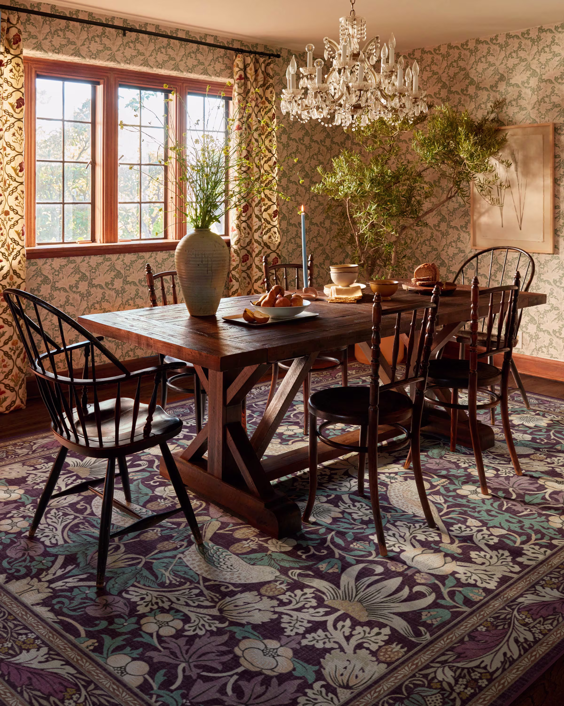 Elegant dining room with a rustic wooden table, surrounded by chairs on a floral rug. A crystal chandelier adds sophistication, and sunlight filters in through a patterned window, creating a warm and inviting atmosphere.