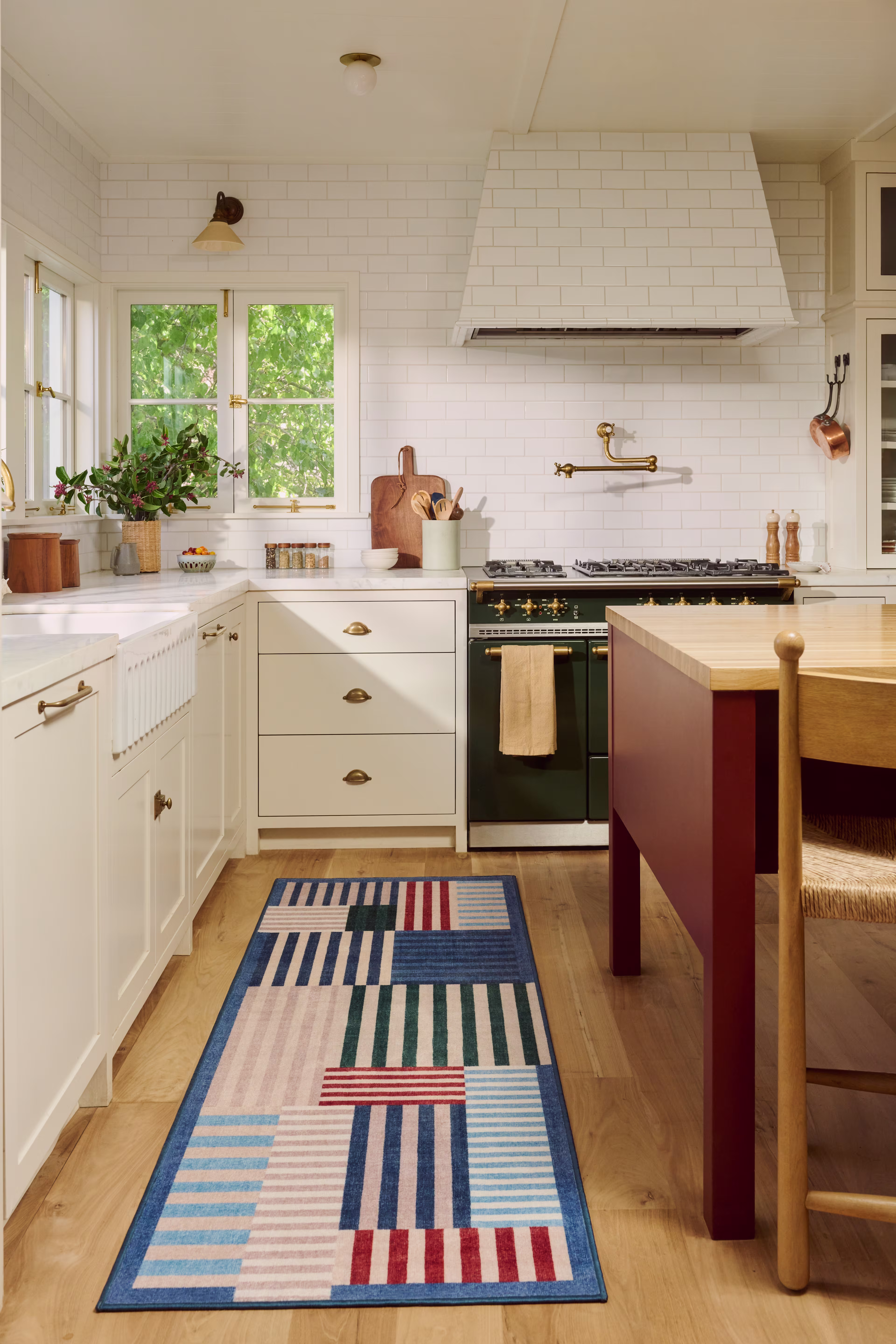 A bright coastal kitchen with white cabinets, a farmhouse sink, a black range, brass fixtures, and a colorful geometric runner rug on light wood floors.