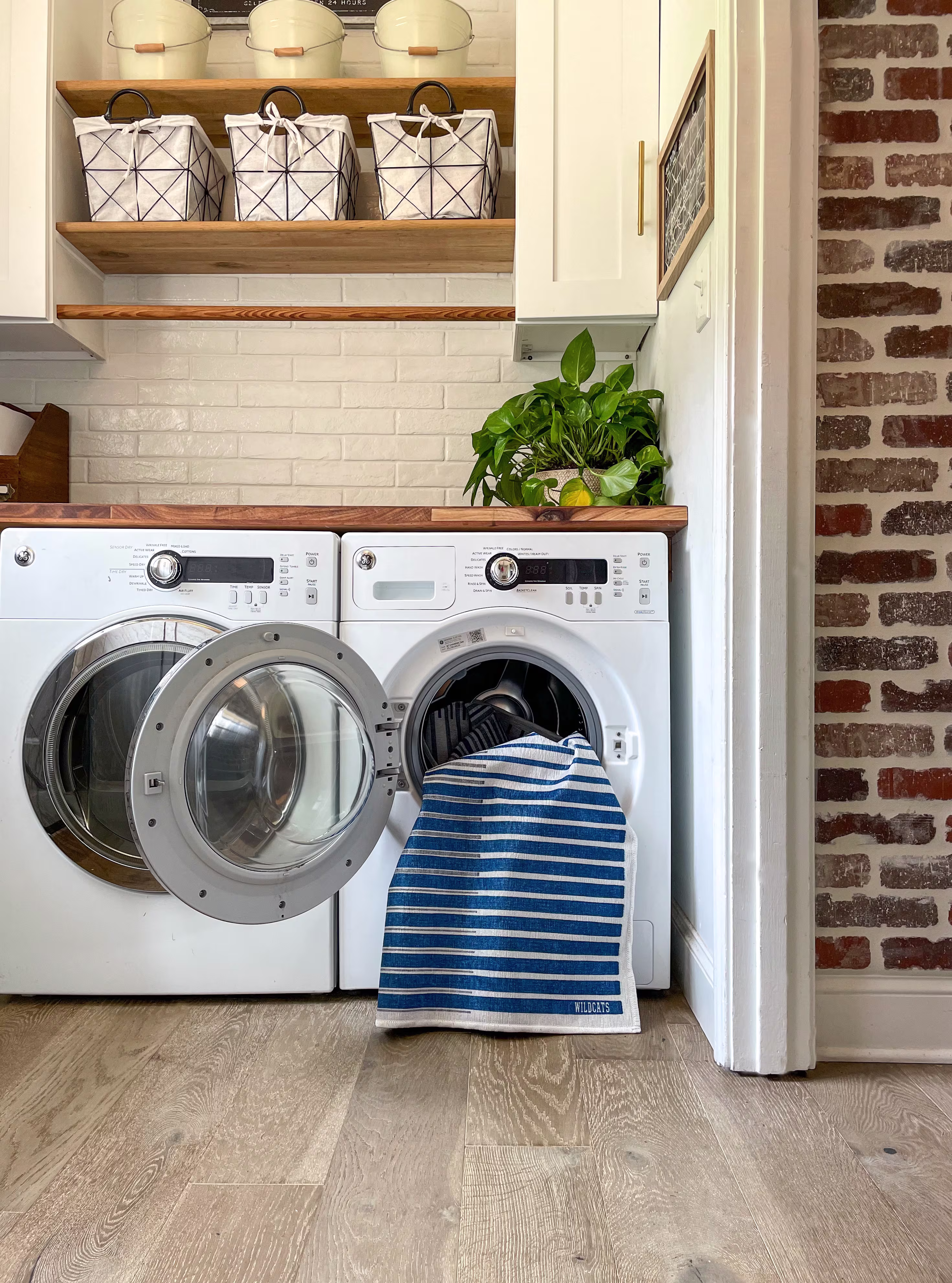 A blue and white striped washable bath rug being pulled out of a front-loading washing machine in a rustic farmhouse laundry room with white subway tile, open wooden shelves, and exposed brick.