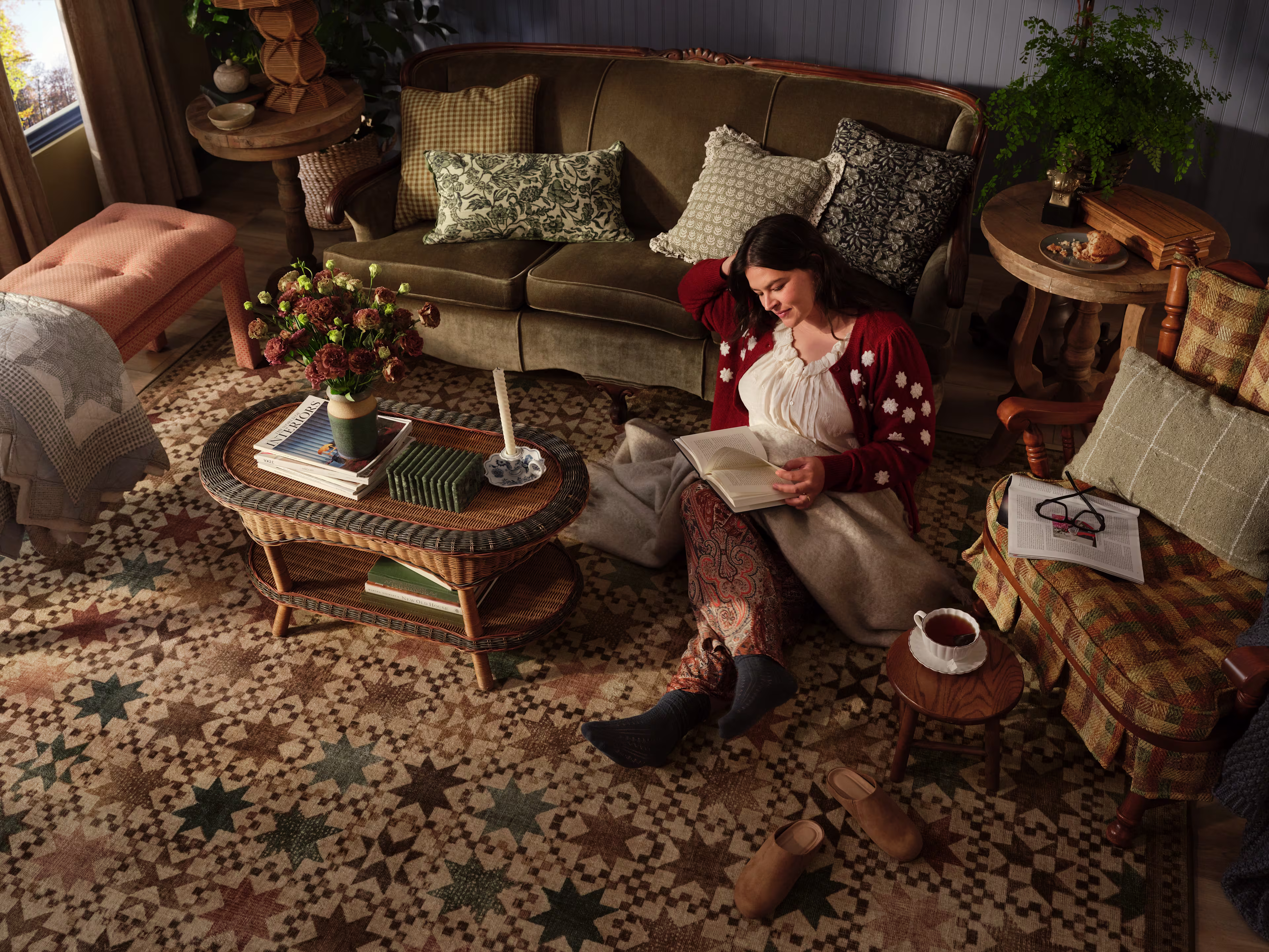 Overhead view of a cozy vintage-style living room featuring a patterned area rug in warm tones, a woman reading on the floor, a velvet sofa, wicker coffee table, and layered textiles creating a warm, inviting interior.