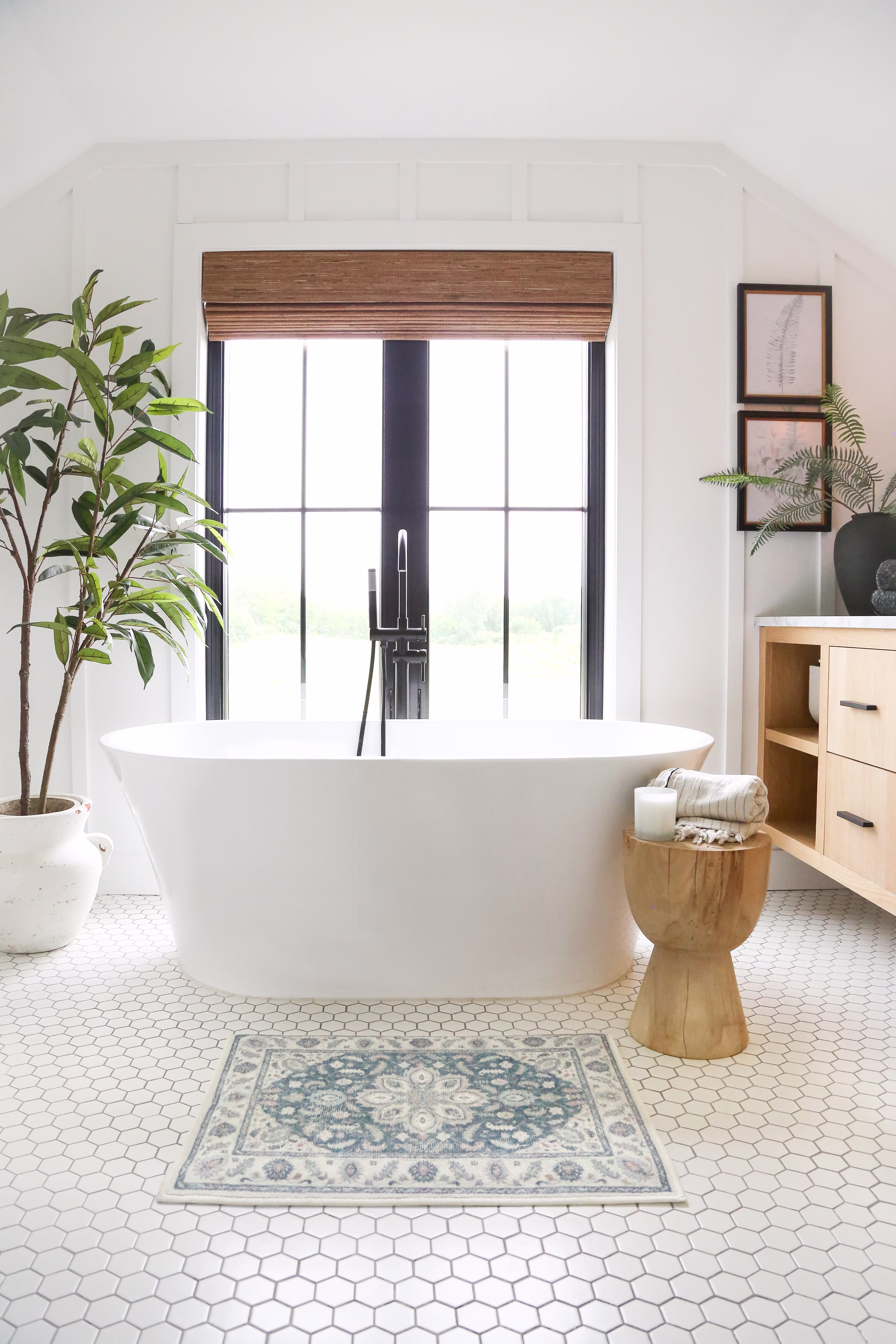 Bright bathroom with a modern white bathtub, wooden stool holding candles and a towel. Large window, indoor plant, and elegant decor create a serene ambiance.