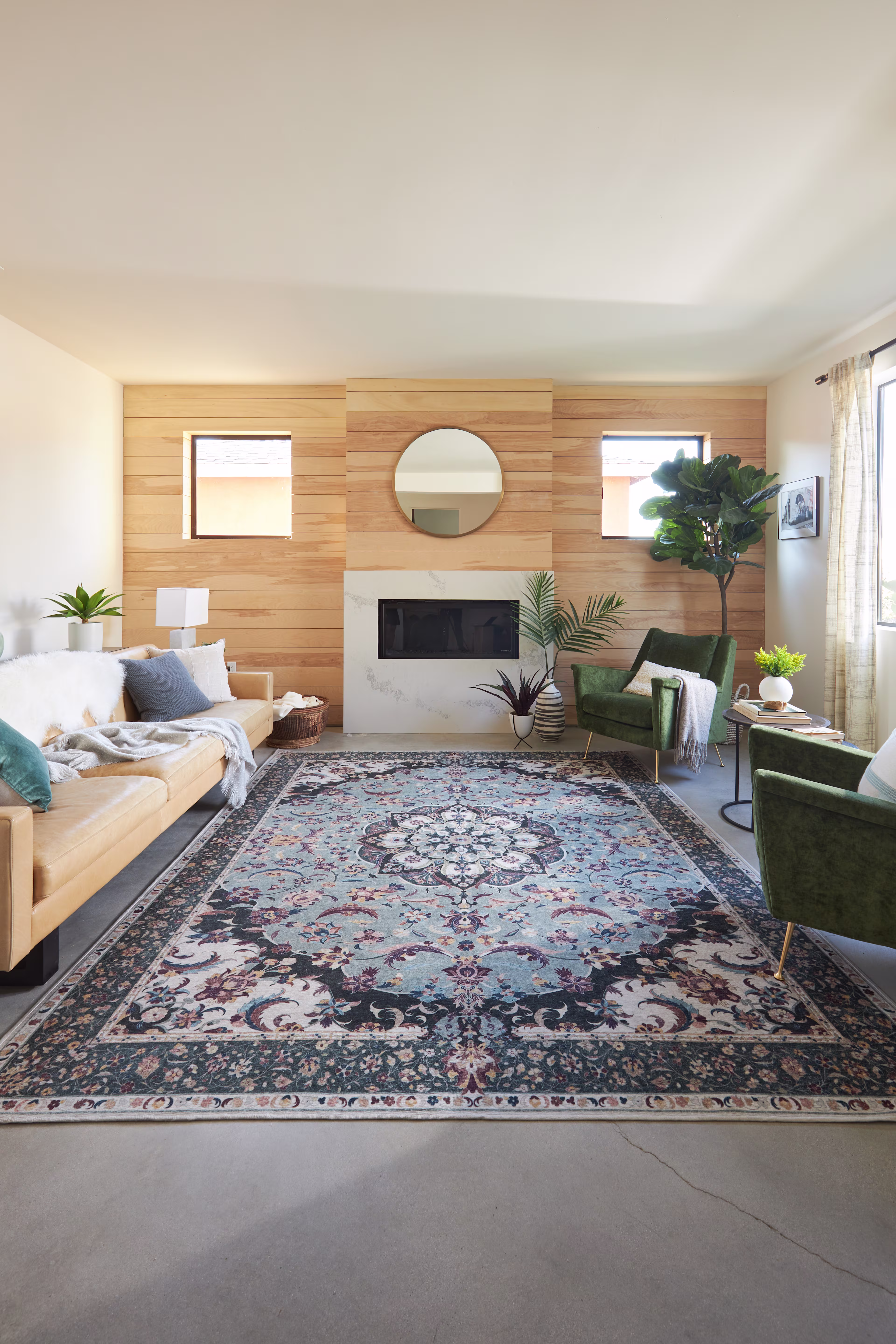 Modern living room with a wooden accent wall, a central fireplace, and a round mirror. Two green chairs face a beige sofa. A large ornate rug adds elegance.