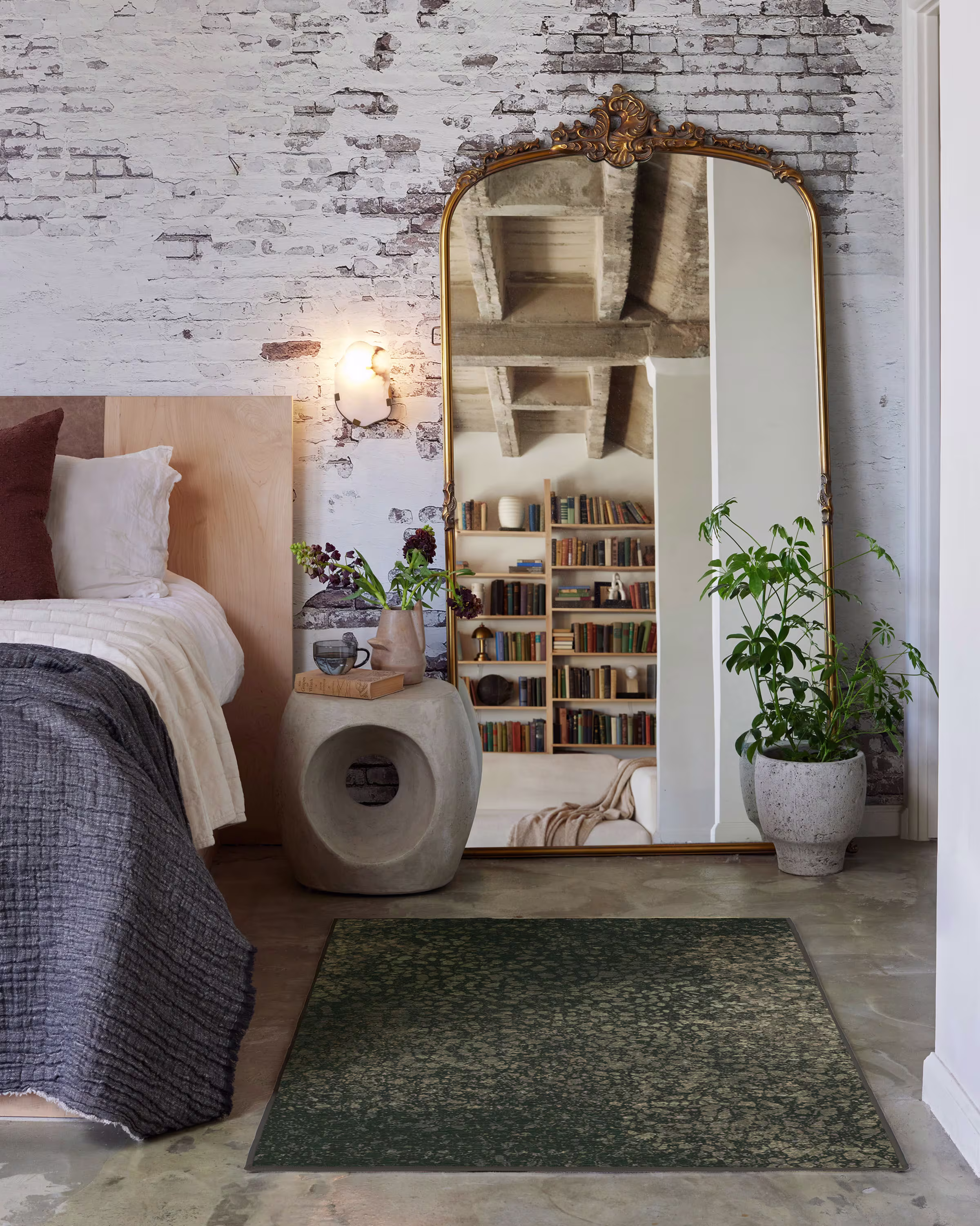 A beautifully styled industrial bedroom featuring a rug from the Architectural Digest collection under a bed. The dark green rug grounds the space, contrasting elegantly with concrete floors, an exposed brick wall, and a large, ornate vintage gold floor mirror.