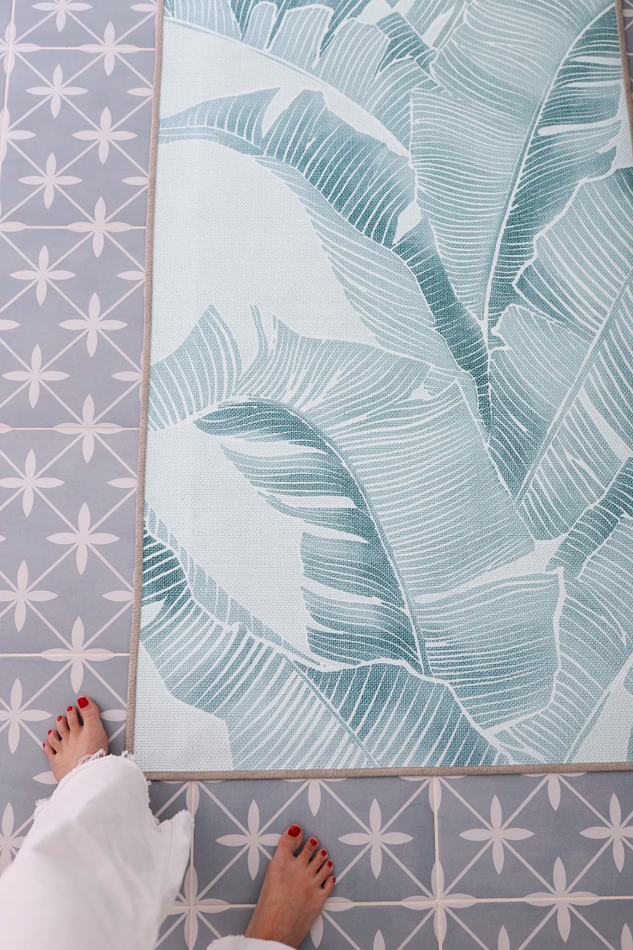 A top-down, POV shot of a person standing next to a Ruggable Gray Malin Banana Leaf Soft Blue bathroom runner rug on a grey and white geometric patterned tile floor.