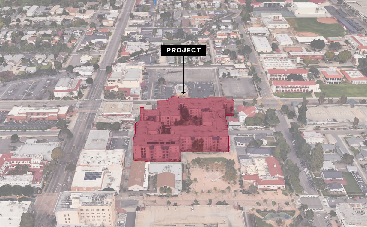A slightly tilted aerial view of an urban block, with a large apartment building area shaded in semi-transparent red at the center. A black label above it reads “PROJECT,” with a thin black arrow pointing down at the highlighted building footprint. Surrounding streets, residential houses, and commercial rooftops appear beyond the shaded area, and tree-lined sidewalks and parking lots frame the block. In the distance, additional rooftops and small urban greenspace are visible.