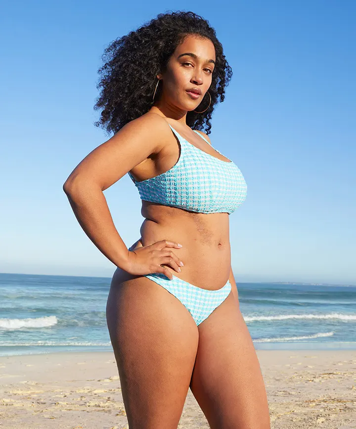 A woman with curly hair stands on the beach wearing a blue checkered bikini, with ocean waves in the background.