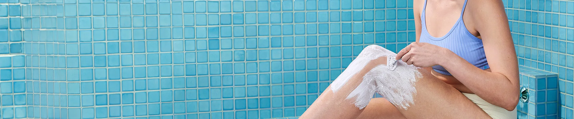 Girl sitting on a bath tub in a blue bathroom shaving her leg