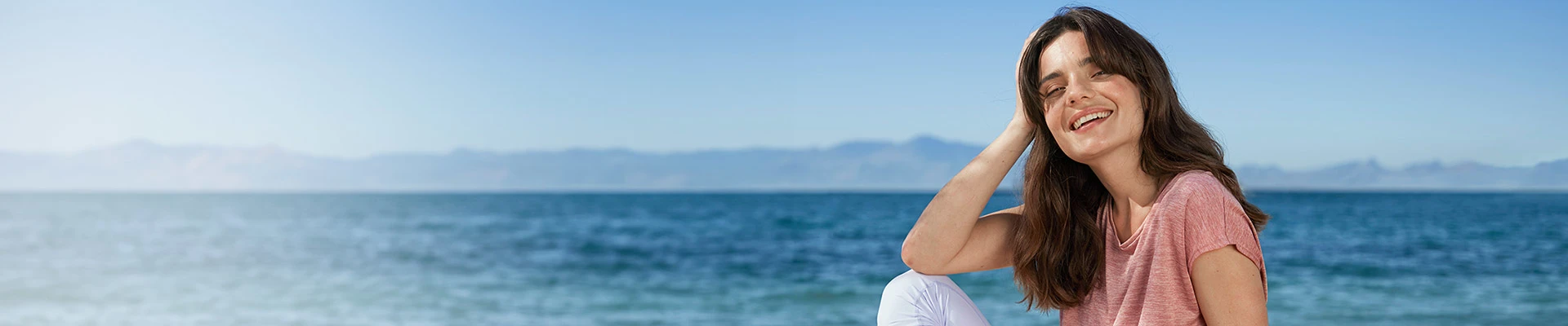 A woman sitting on the beach, smiling, wearing a pink shirt and white pants with water and mountains in the background.