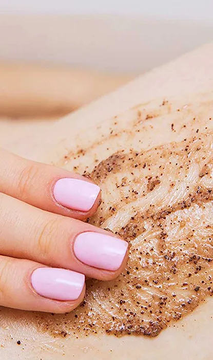 Hand with pink nails applying a brown scrub to skin, showcasing a beauty treatment for exfoliation and skin care benefits.