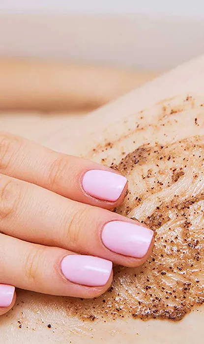 A close-up of a hand with pink nails applying a body scrub on the skin, showcasing smooth and exfoliated texture.