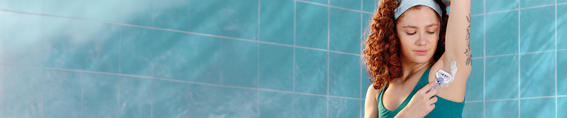 Woman with curly red hair with a headband sitting on a bath tub, shaving her armpit