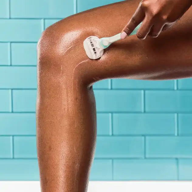 Woman using a razor to shave a lower leg against a tiled background