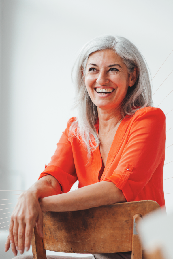 A woman wearing a tomato red shirt sits backward on a chair, smiling.