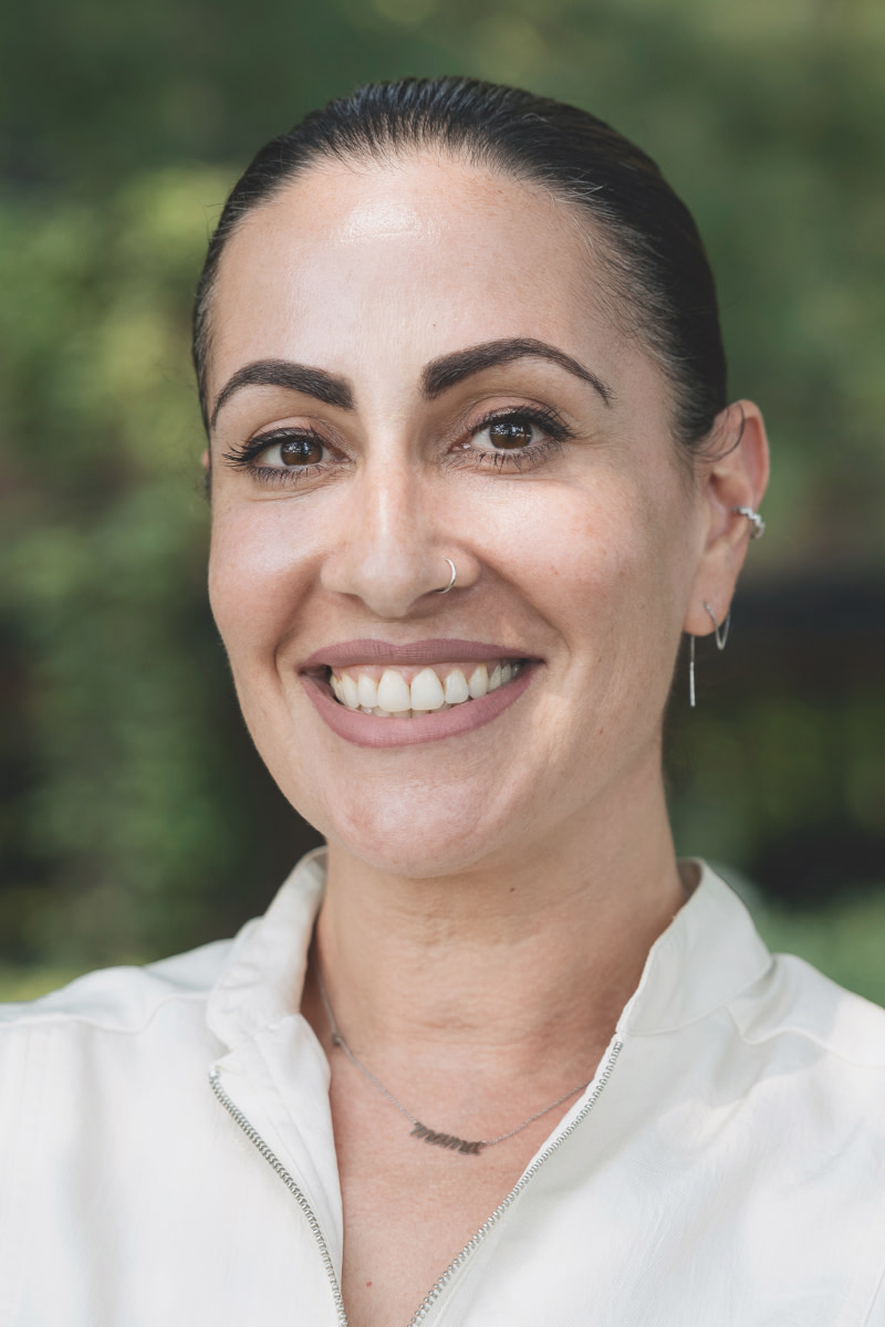 Portrait of a woman smiling. She is wearing a white shirt and standing in front of a tree outdoors.