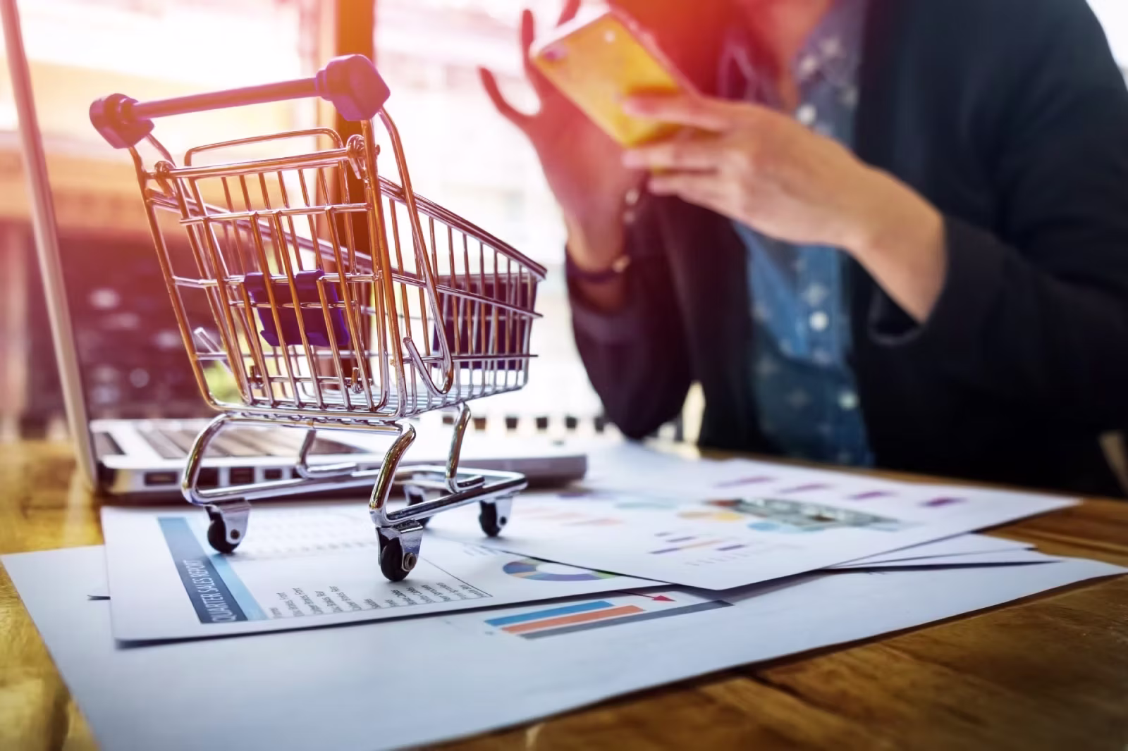 Mini shopping cart placed on financial reports with a person using a smartphone in the background
