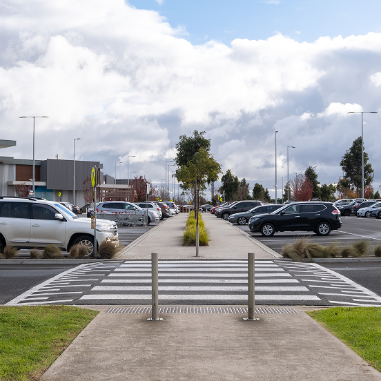 Outdoor shopping center parking lot with pedestrian crossing, rows of parked cars, and cloudy blue sky overhead.