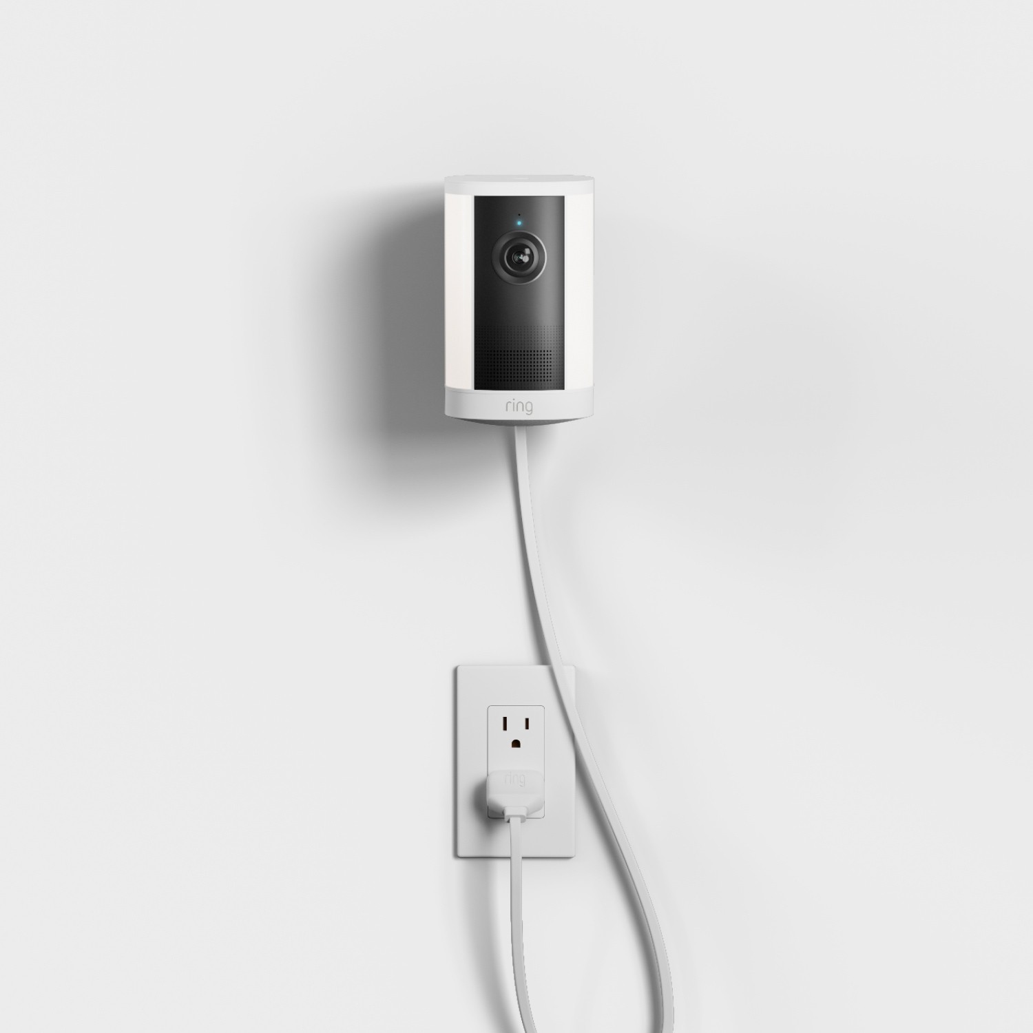 Spotlight Cam 4K Plus - Plug-In:White Ring indoor camera mounted on a wall, powered by a long white cord plugged into a nearby outlet on a plain white background.