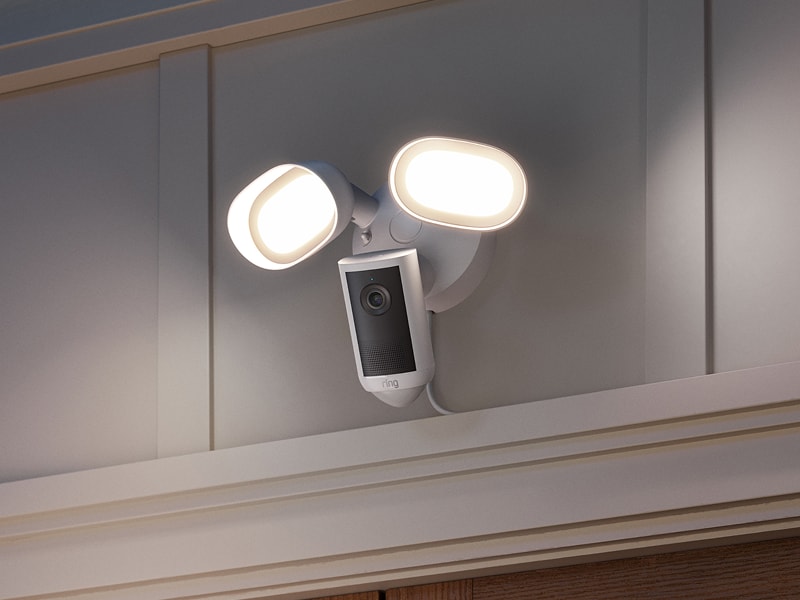 Outdoor wall-mounted floodlight with integrated camera, two bright LED lamps glowing on a house exterior at dusk.