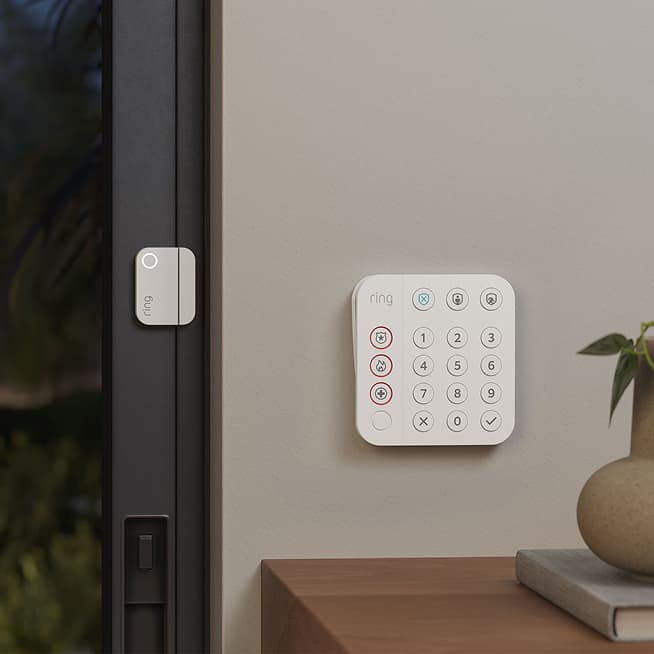 Ring keypad on a wall beside a sliding door, with a small contact sensor on the door frame and a plant and book on a nearby table.