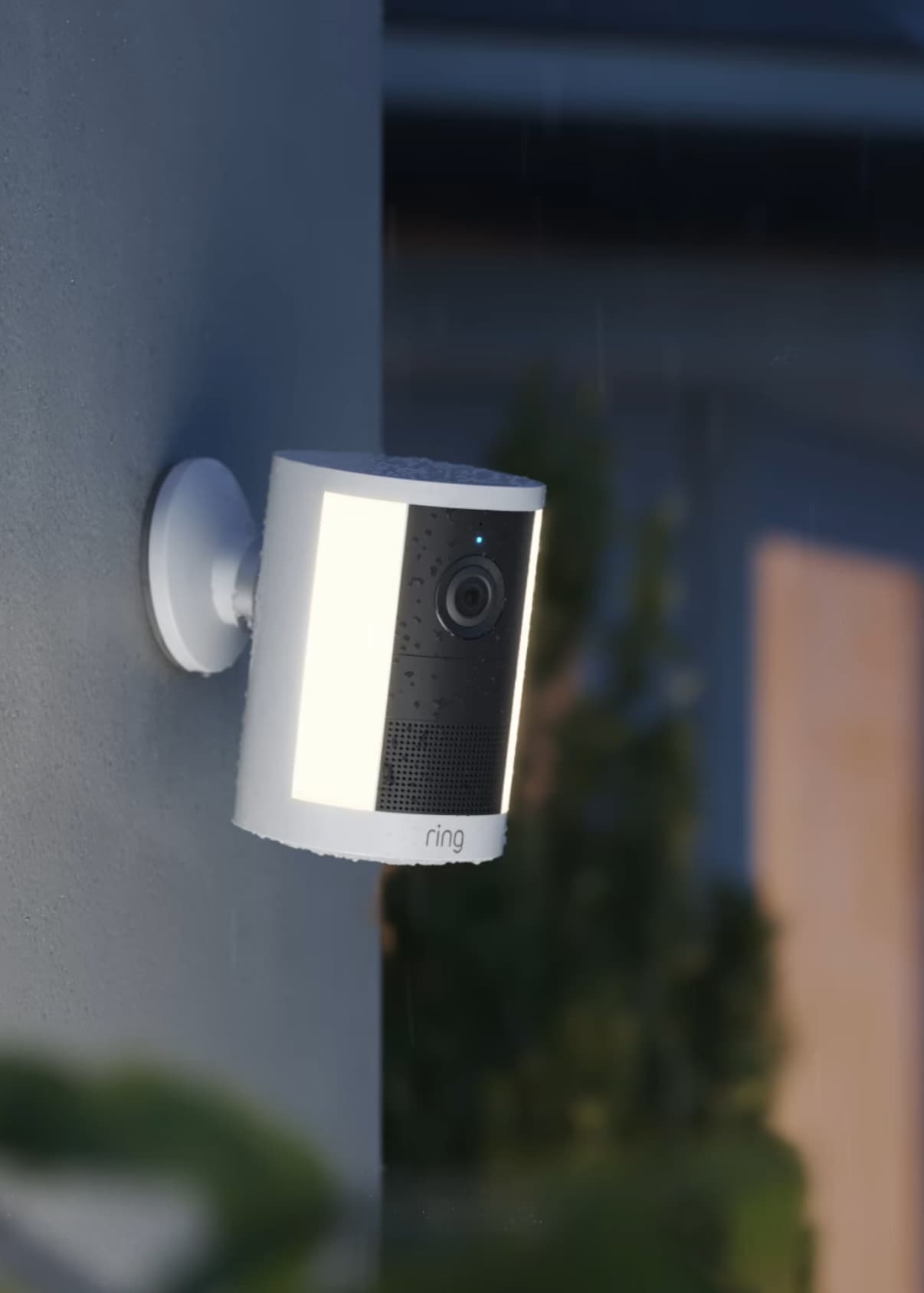 Spotlight Cam (2nd Gen) mounted on an outside wall, raining at dusk with warm window glow.