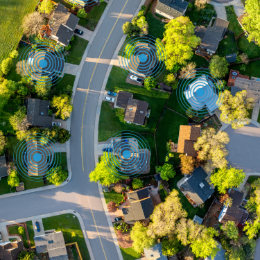 Aerial view of a suburban street with trees and homes, featuring blue concentric signal icons over several yards indicating device zones.