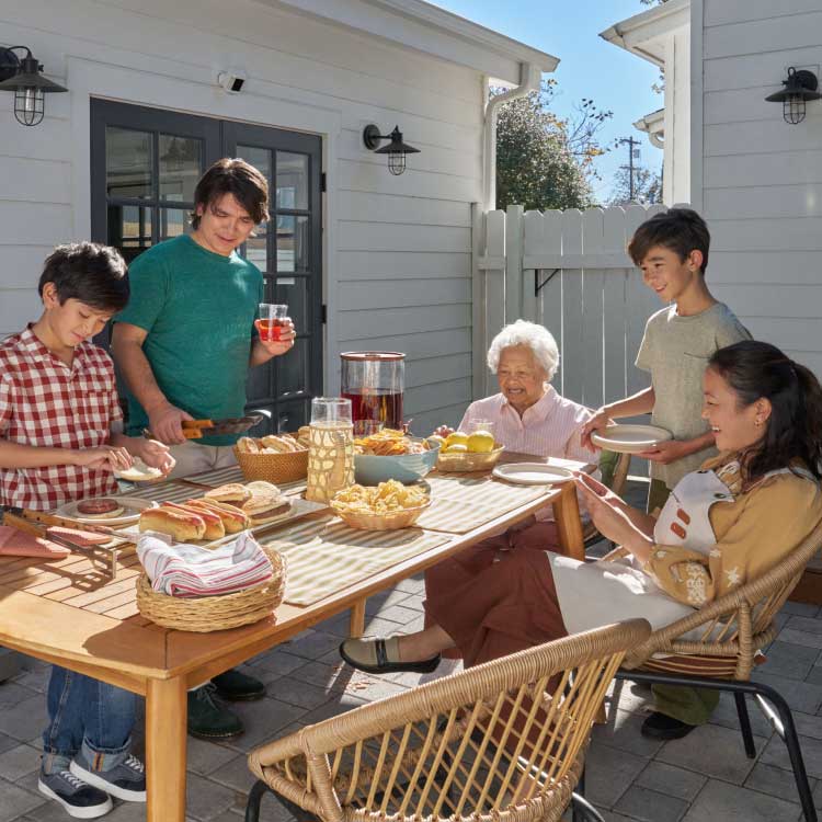 Family enjoying a sunny backyard cookout by a grill, preparing burgers and snacks around a table with drinks and fruit.