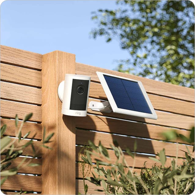 Outdoor security camera with attached solar panel mounted on a wooden fence, with leafy greenery and blue sky in the background.