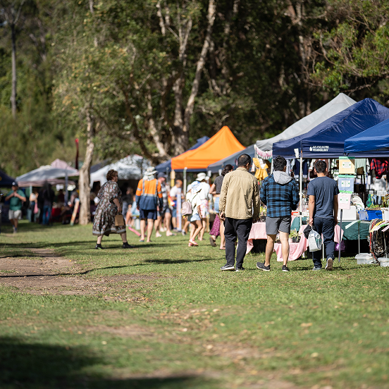 Outdoor market with colorful vendor tents in a grassy park area with people browsing stalls under tall trees.