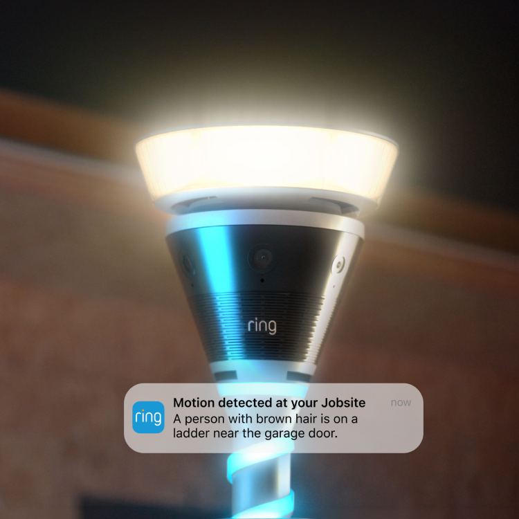 Close-up of a Ring outdoor floodlight camera glowing in warm light, with a smartphone alert overlay about motion near a garage door.