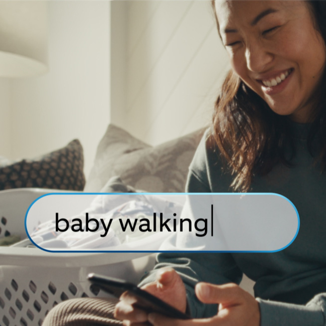 Smiling woman on couch holding smartphone, next to laundry basket. A Ring IQ search bar overlay reads 'baby walking.'