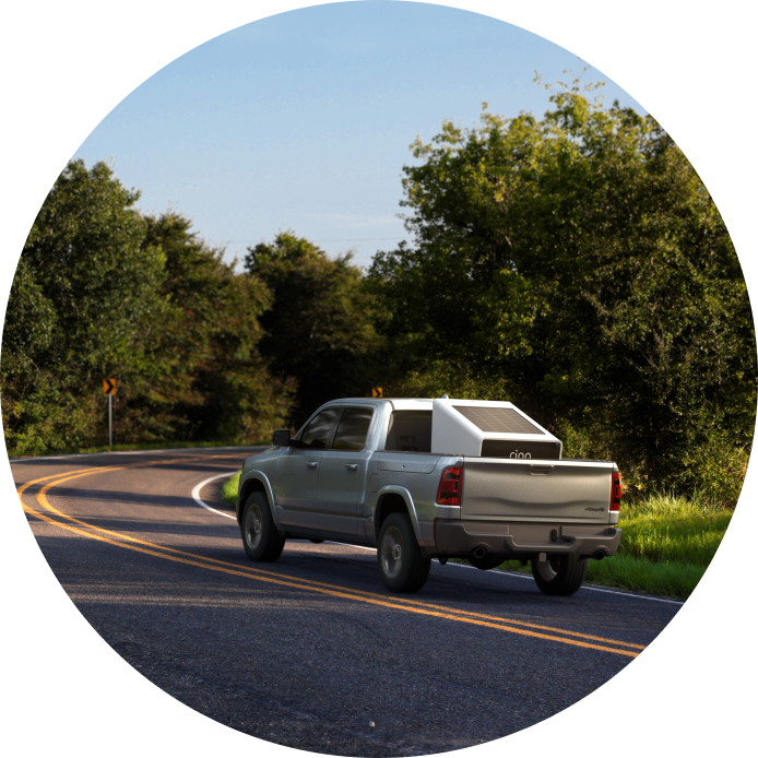Silver pickup with a Ring security mobile tower equipment unit in the bed drives along a winding rural road bordered by green trees under a clear sky.