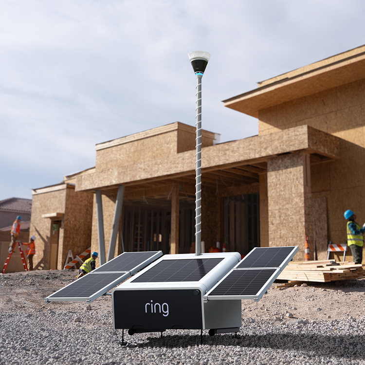 Solar-powered Ring mobile security tower unit with tall mast set on gravel at a wood-frame construction site, workers in hard hats in the background.