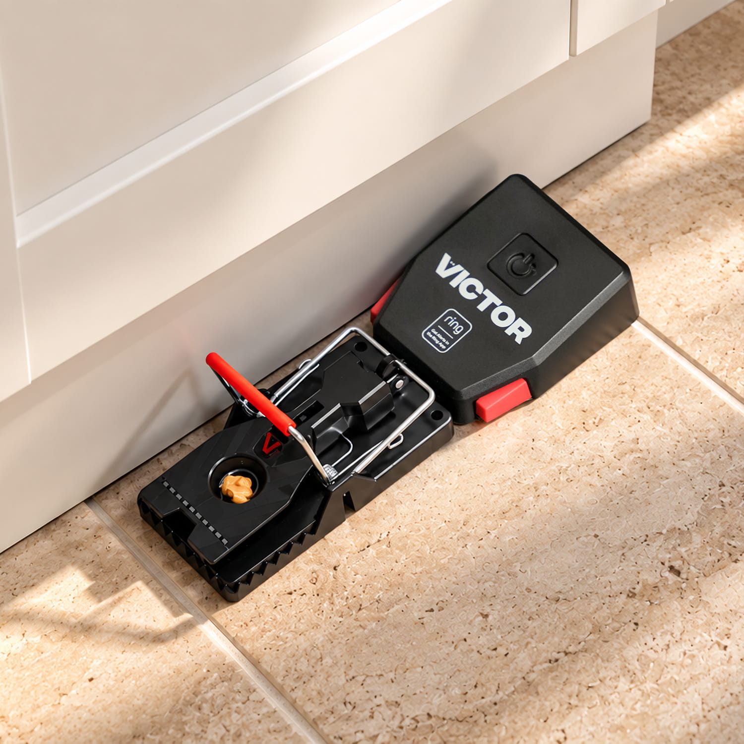 Victor® Smart Mouse Trap, Indoor Snap Trap - Black Victor electronic mouse trap placed along a kitchen baseboard on tan tile, baited and ready with red safety latch visible.