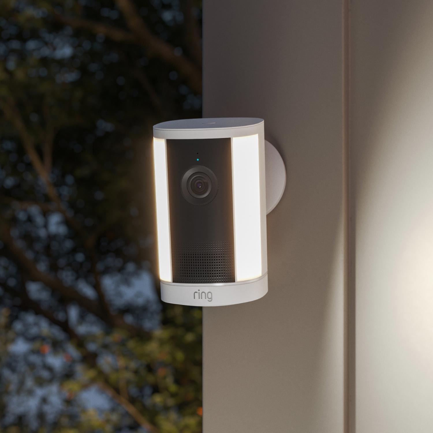 Spotlight Cam 4K Plus - Battery:Wall-mounted Ring camera with dual LED light strips glowing at dusk, set against a blurred background of trees.