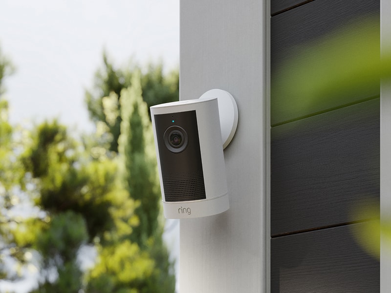 White Ring outdoor camera mounted on a modern house wall, with trees and soft daylight in the background.