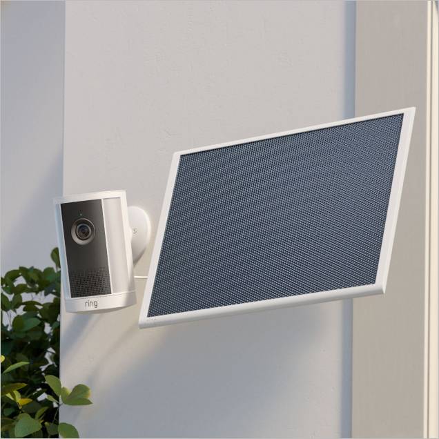White outdoor Ring camera mounted on a wall next to a tilted rectangular solar panel, with green leaves in the corner.
