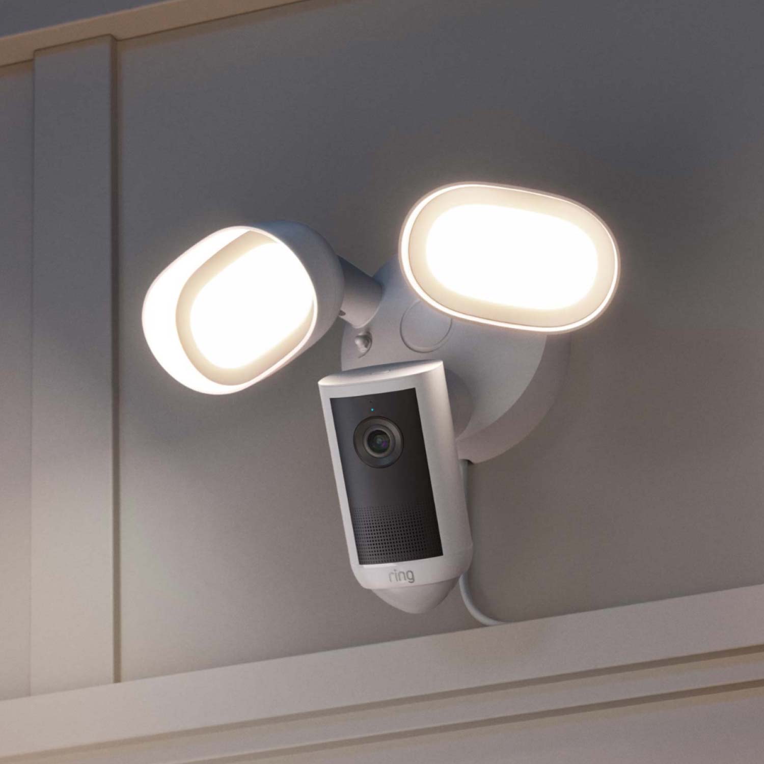 Floodlight Cam 4K Plus - Plug-In:Outdoor wall-mounted Ring floodlight camera with two bright LED lights turned on, attached near a roof eave at dusk.
